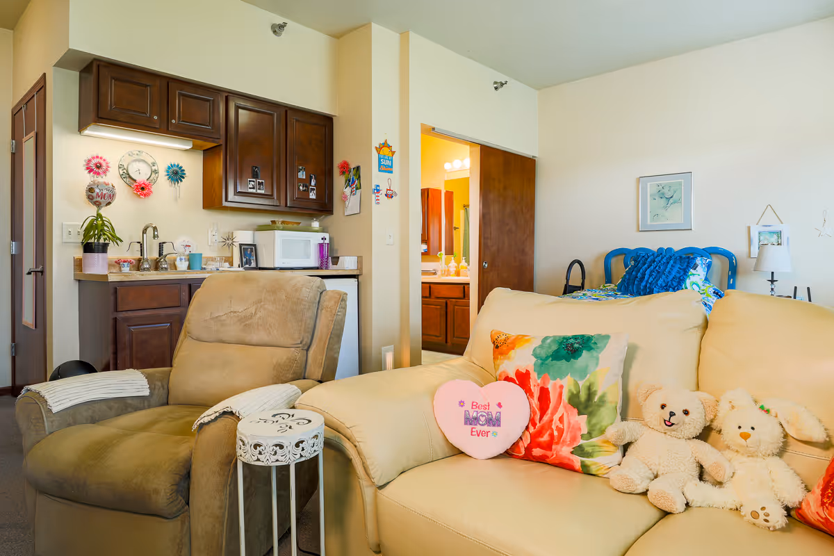 A cozy assisted-living apartment interior showing a recliner and sofa with colorful pillows and stuffed animals, a small kitchenette with cabinets and microwave, and a doorway to a bathroom.