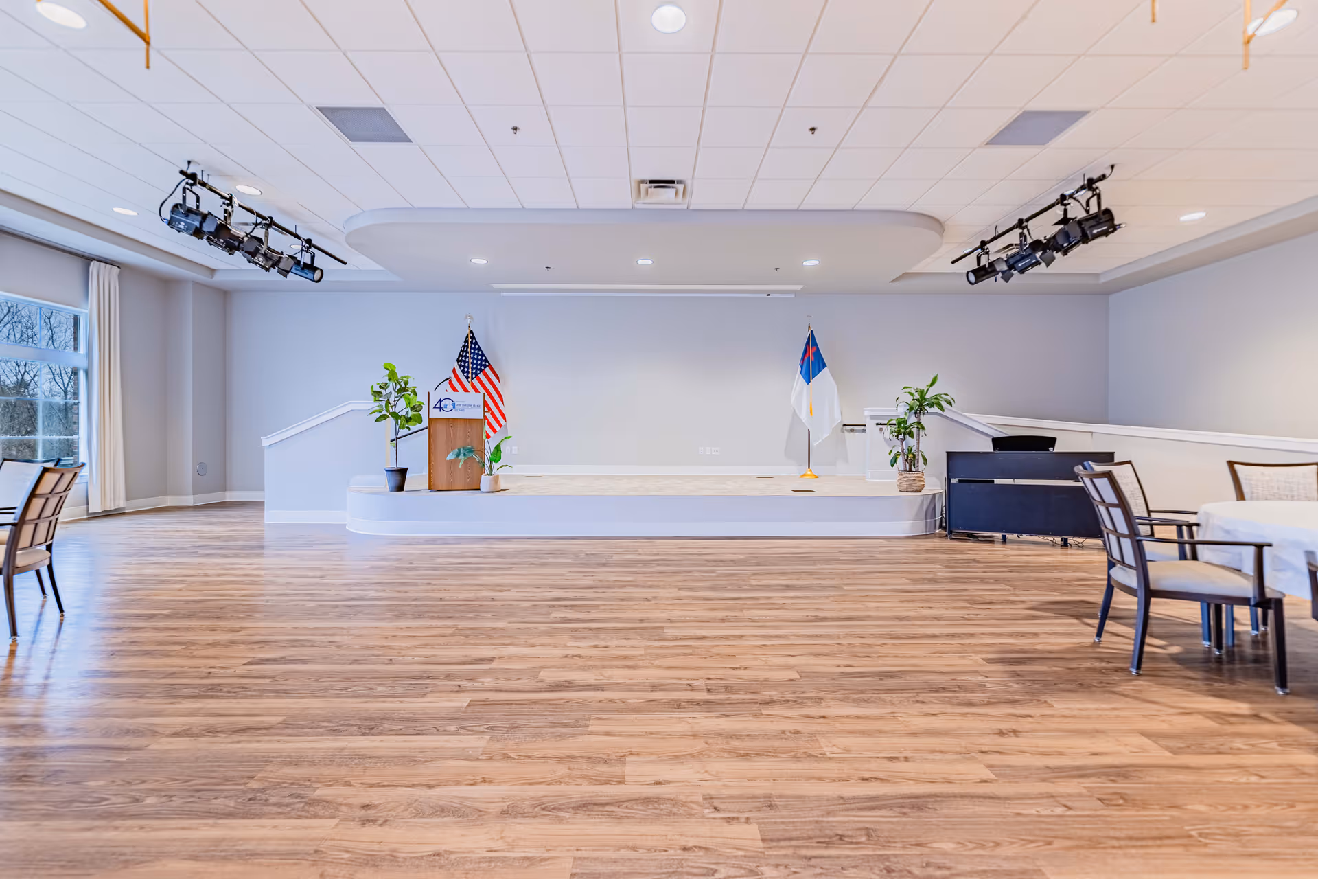 Large empty activity room with a small raised stage and podium, flags, round tables and chairs on wood flooring.