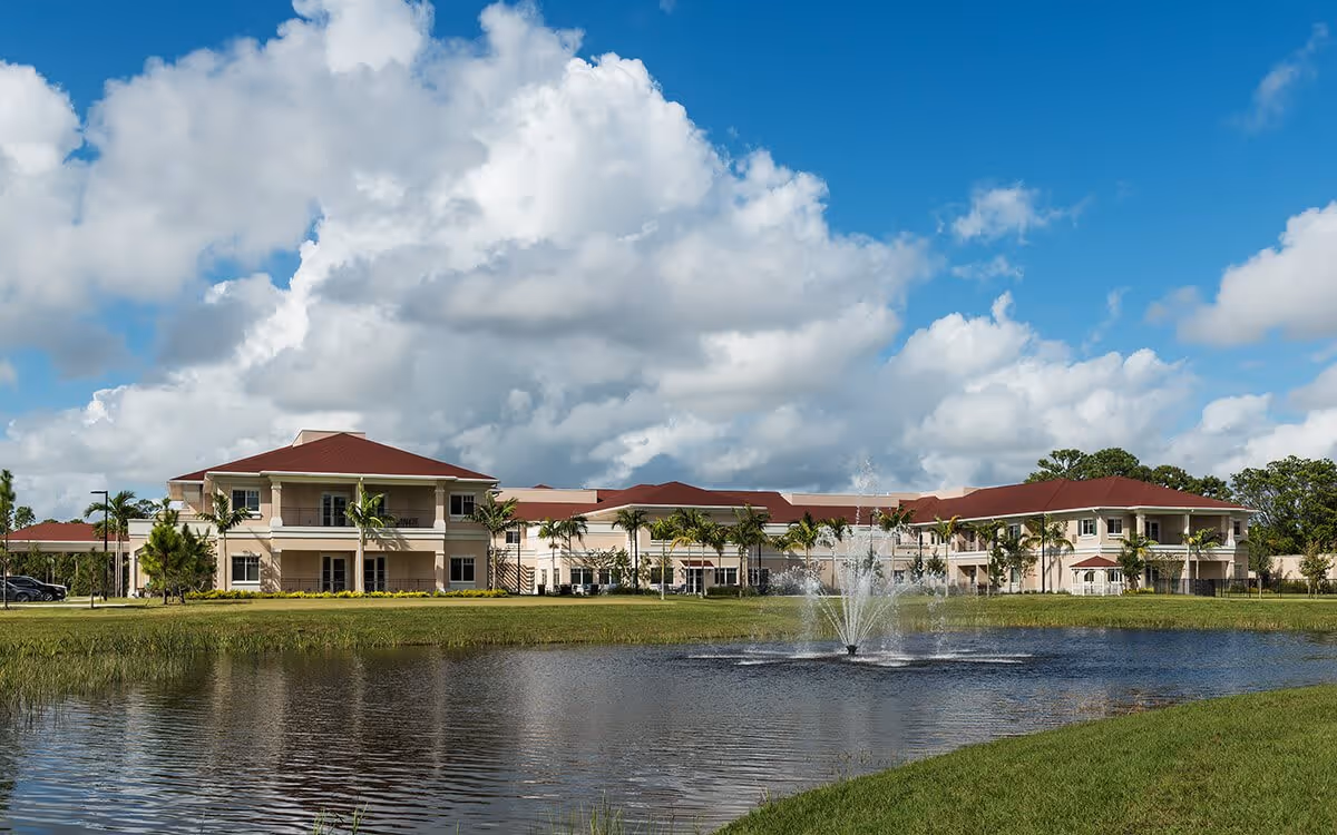 Exterior view of a senior living facility with beige buildings and red roofs, surrounded by palm trees and greenery. In the foreground, there is a pond with a water fountain spraying water upwards under a partly cloudy blue sky.
