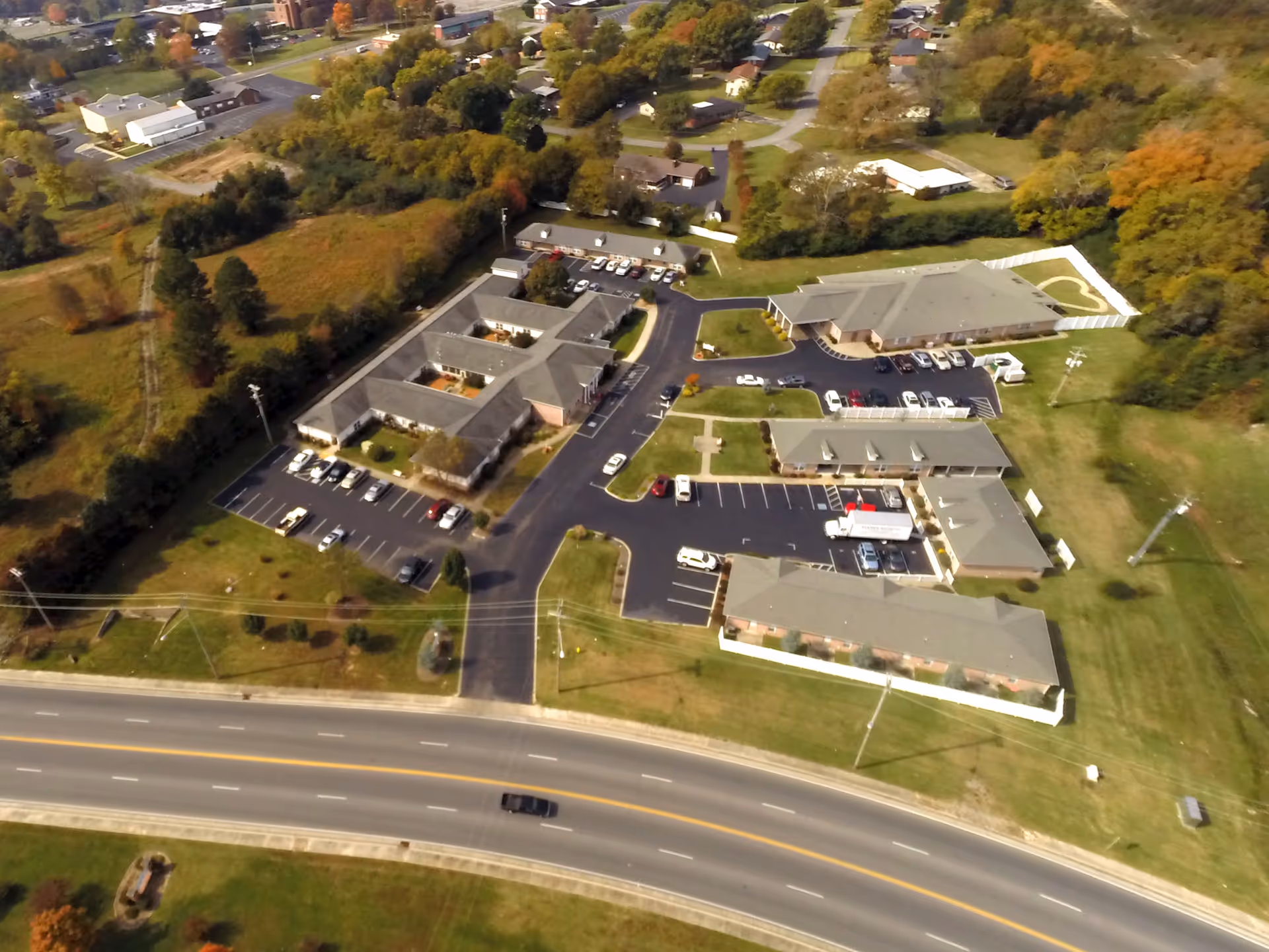 Aerial view of Carriage House Inn facility showing multiple single-story buildings with parking lots, surrounded by green lawns and trees with autumn foliage, adjacent to a main road.