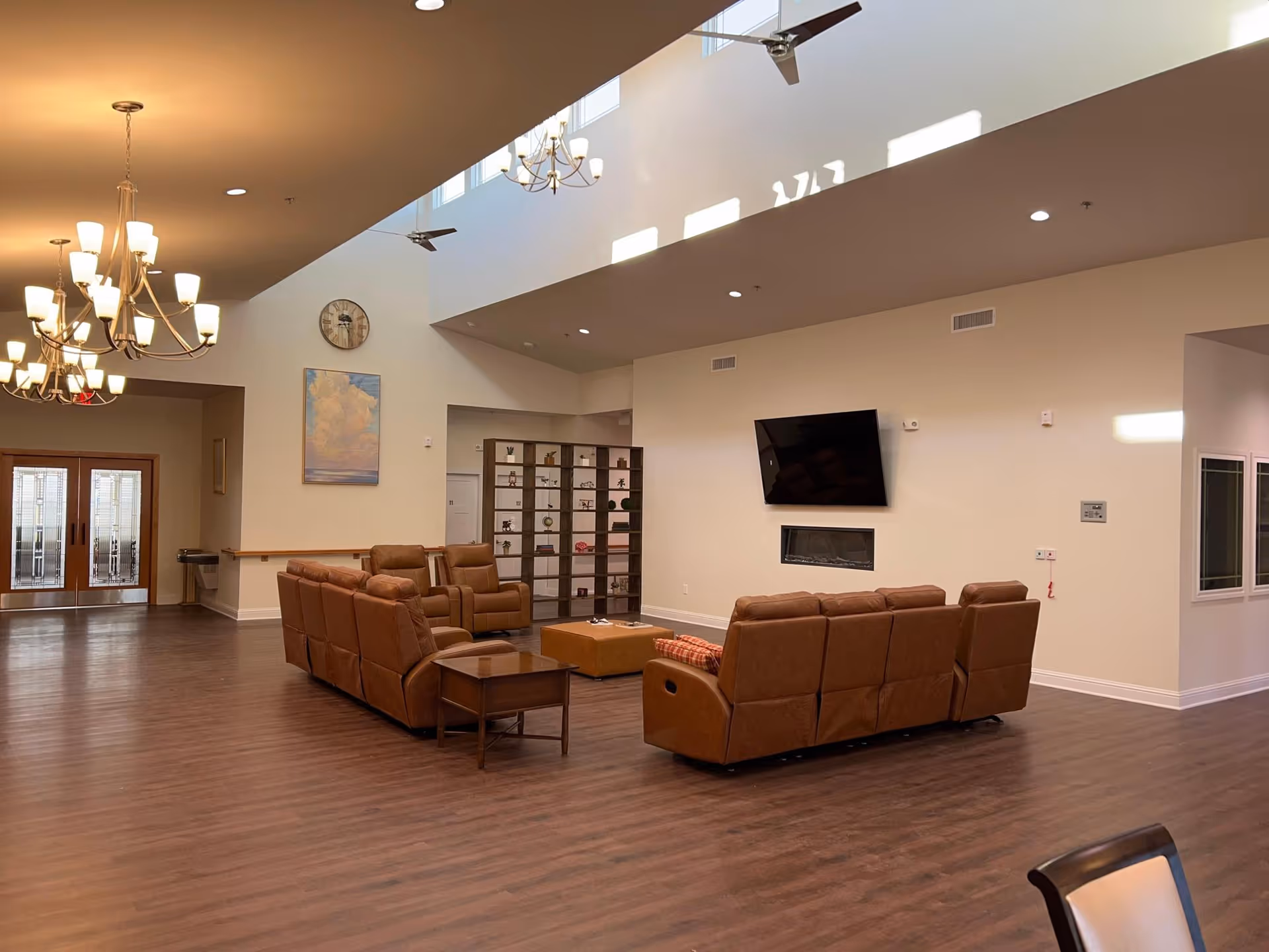 A spacious senior living facility common area with brown leather sofas arranged around a wall-mounted flat screen TV and a modern fireplace. The room features high ceilings with ceiling fans and chandeliers, wood flooring, a large clock on the wall, and a decorative shelving unit. Double glass doors are visible in the background.