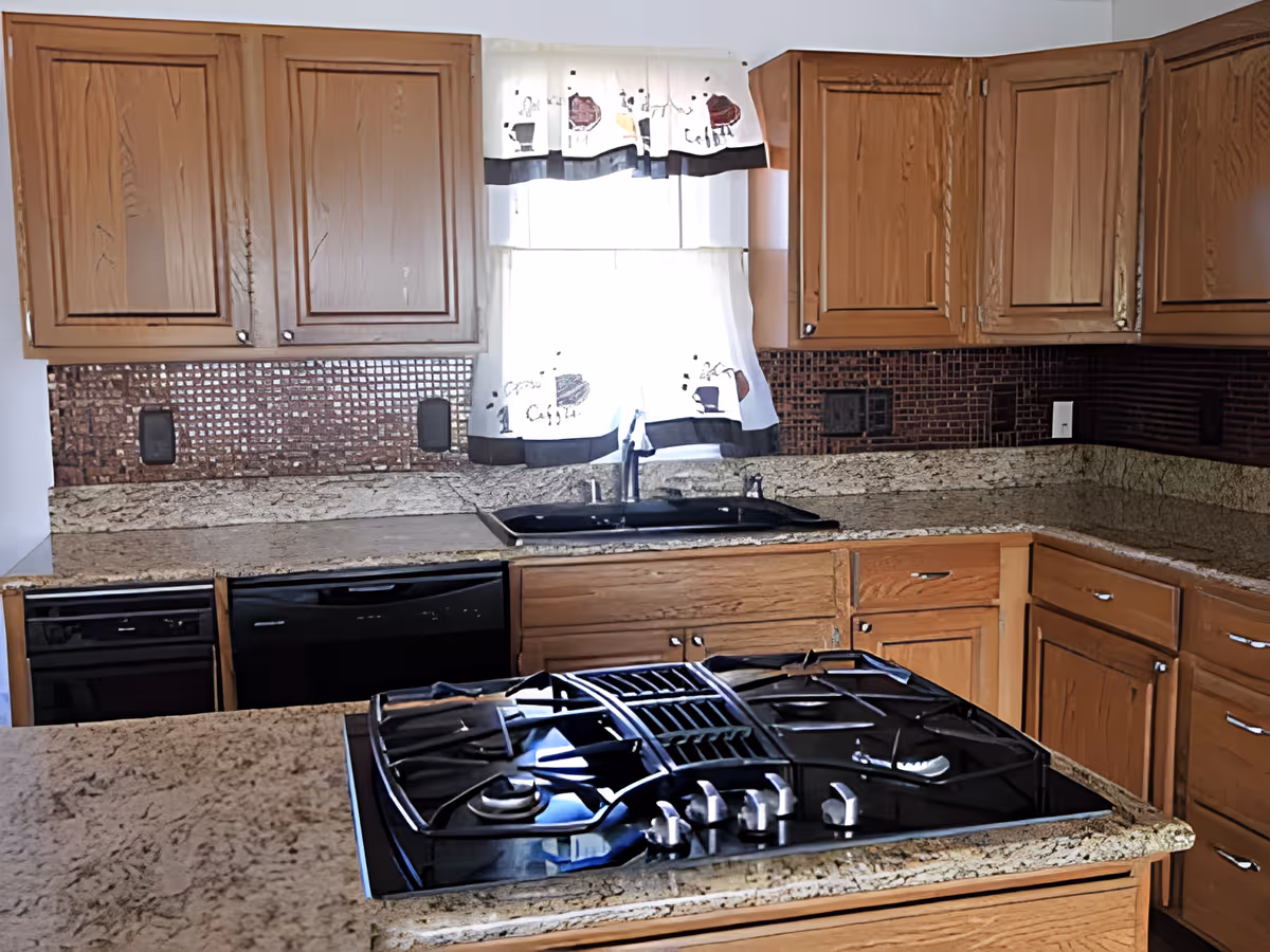 A kitchen with wooden cabinets, granite countertops, a black gas stove on an island, a black dishwasher, and a black sink under a window with white curtains featuring a coffee cup pattern.