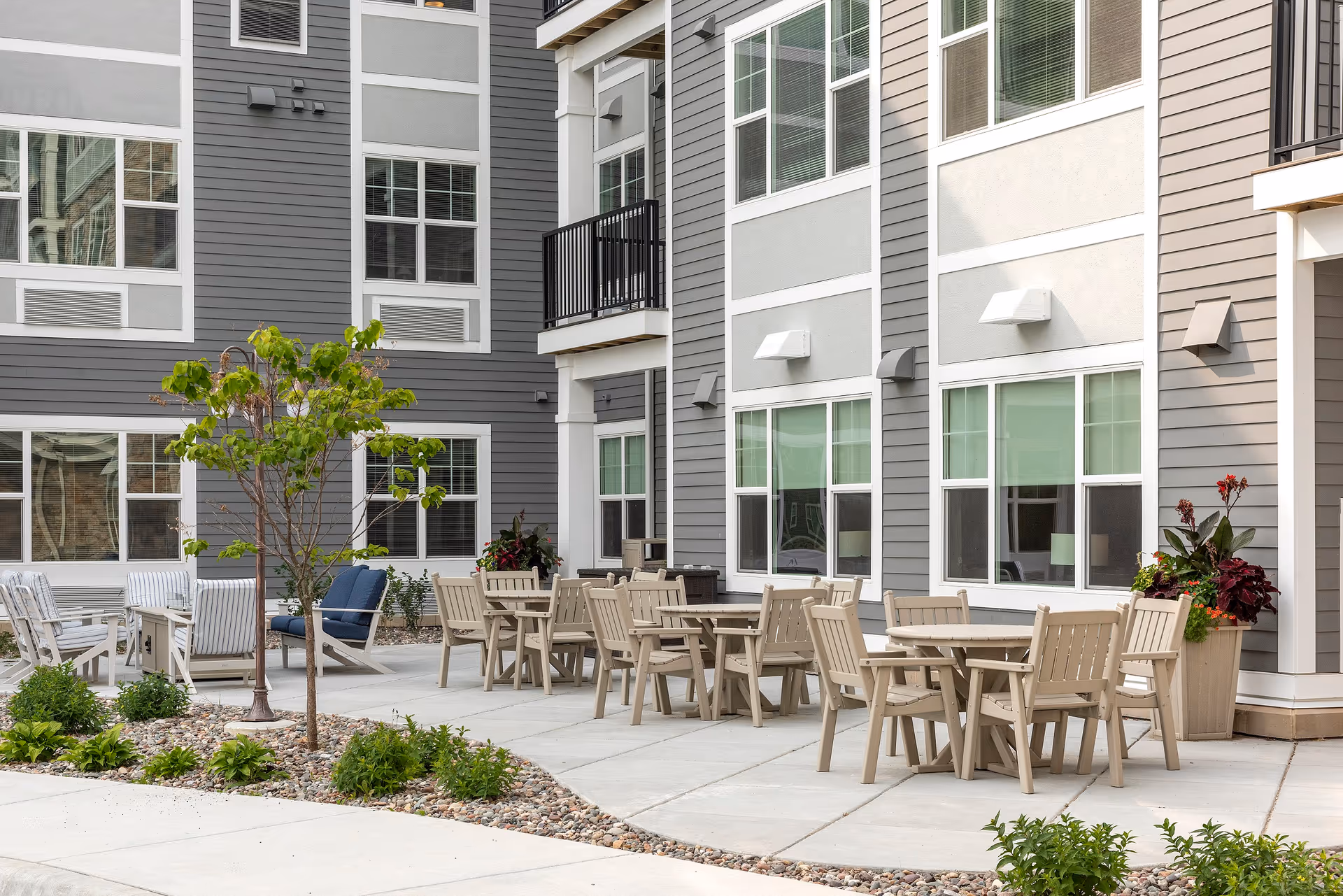 Outdoor patio area at Havenwood of Maple Grove featuring multiple beige wooden tables and chairs arranged on a concrete surface, with a small tree and landscaped plants surrounded by rocks. The building exterior is visible with gray and white siding and several windows.