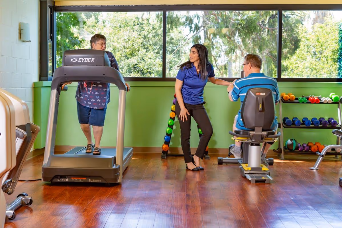 An elderly woman walking on a treadmill and an elderly man using a stationary exercise bike in a fitness room with a female instructor assisting them. The room has large windows showing green trees outside and a rack of colorful dumbbells against a green wall.