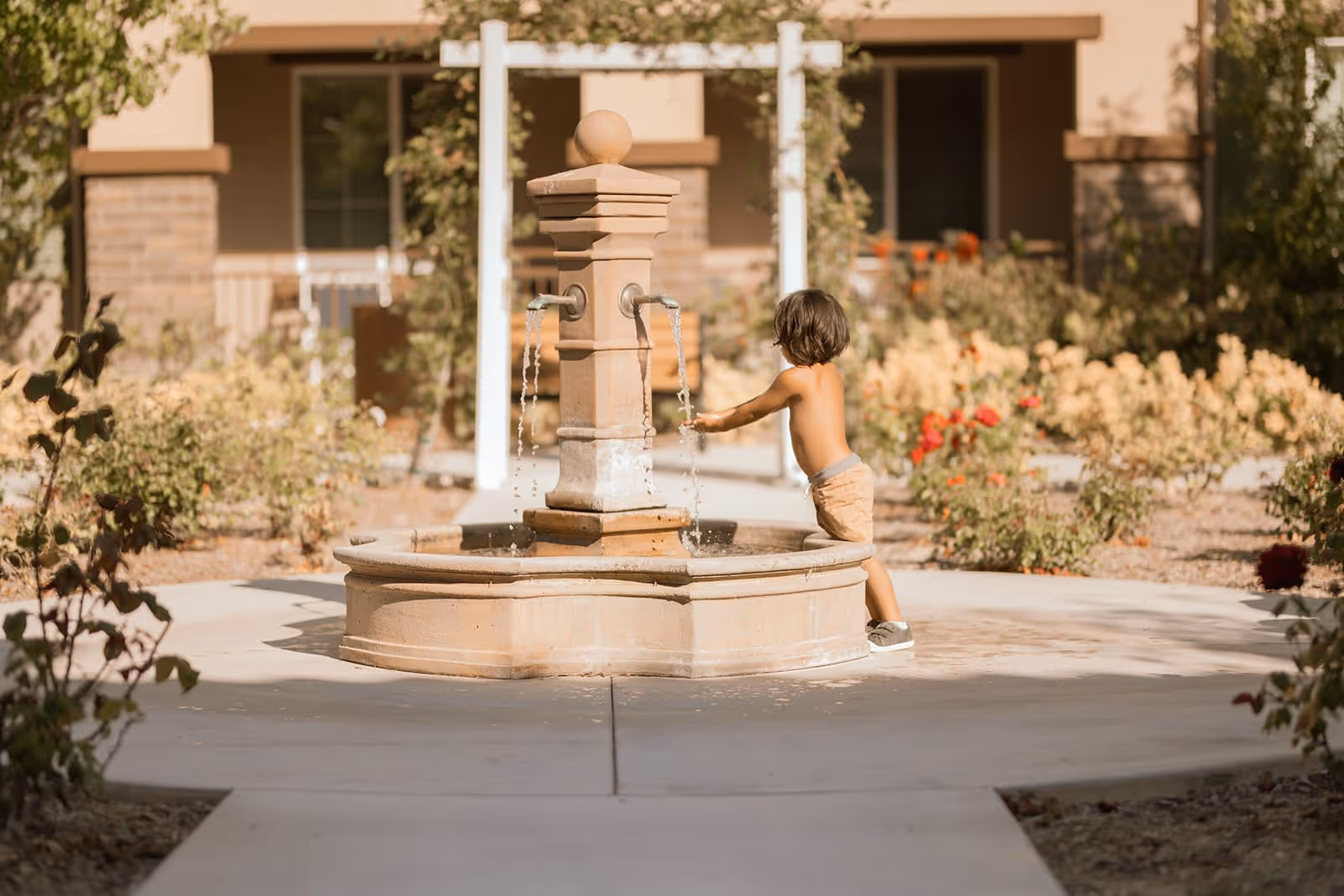 A young child with dark hair, wearing shorts and sneakers, plays with water flowing from a stone fountain in an outdoor garden area with plants and a building in the background.