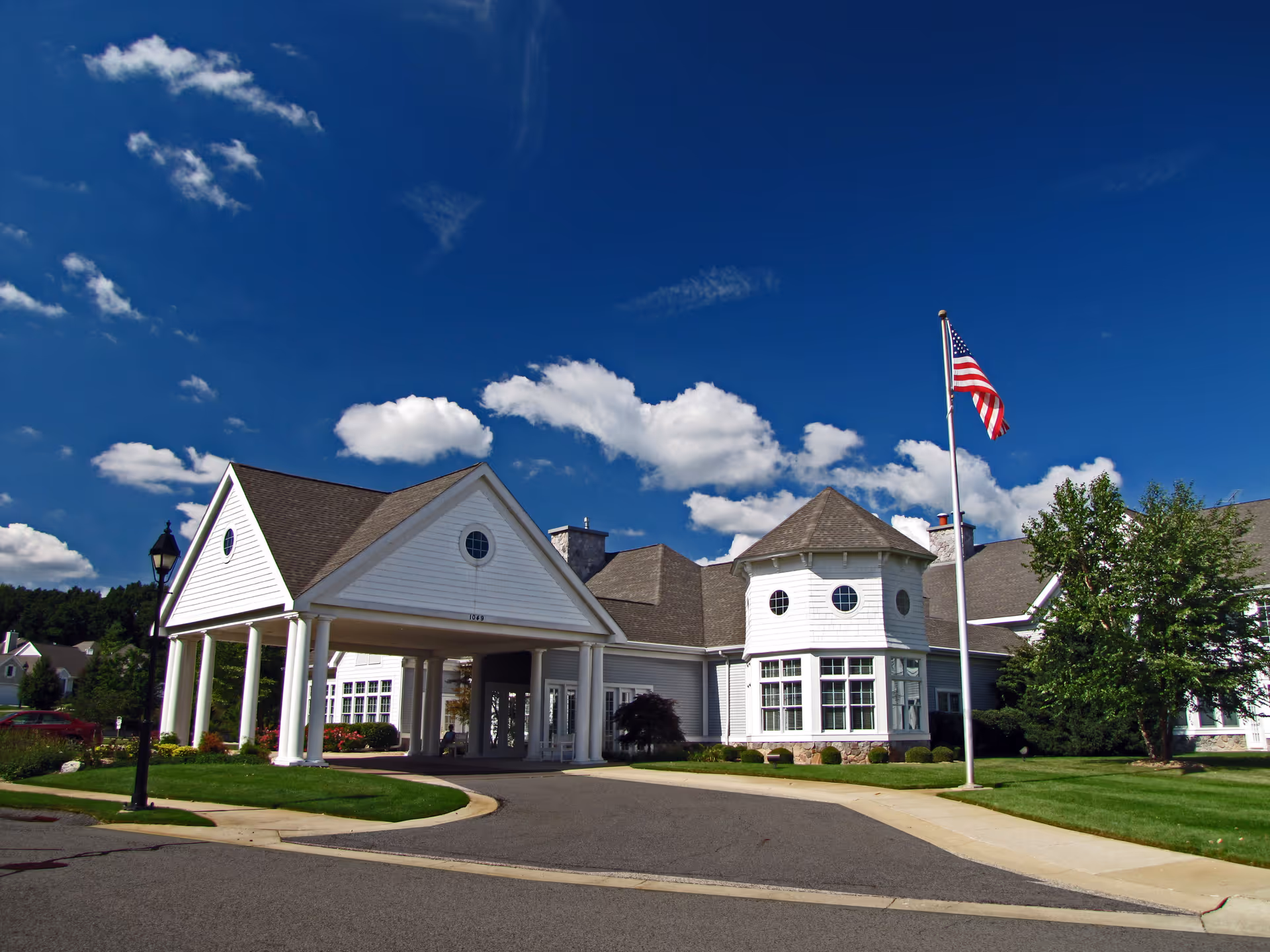 White two-story senior living building with a covered entrance, flagpole flying an American flag, and a bright blue sky.