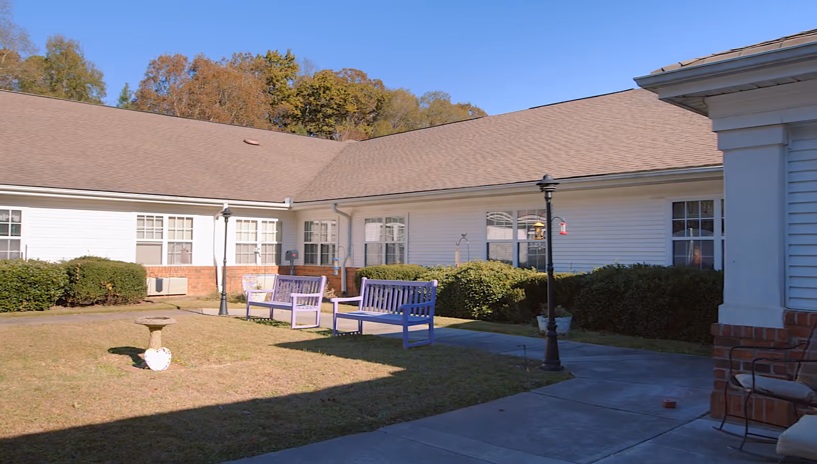 Sunny courtyard with purple benches, lamp posts, a birdbath, and a one-story white senior living building in the background.