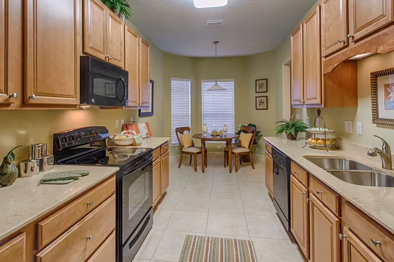 Well-lit kitchen with wooden cabinets, black appliances, a double sink, and a small breakfast table by three windows.