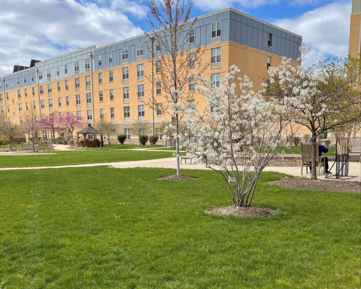 Outdoor view of a senior living facility with a large green lawn, blossoming trees, benches, and a gazebo. The building in the background is a multi-story structure with many windows under a partly cloudy sky.