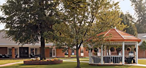 Outdoor view of a senior living facility with a white gazebo surrounded by trees and green grass. In the background, there is a single-story brick building with a gray roof and several windows.