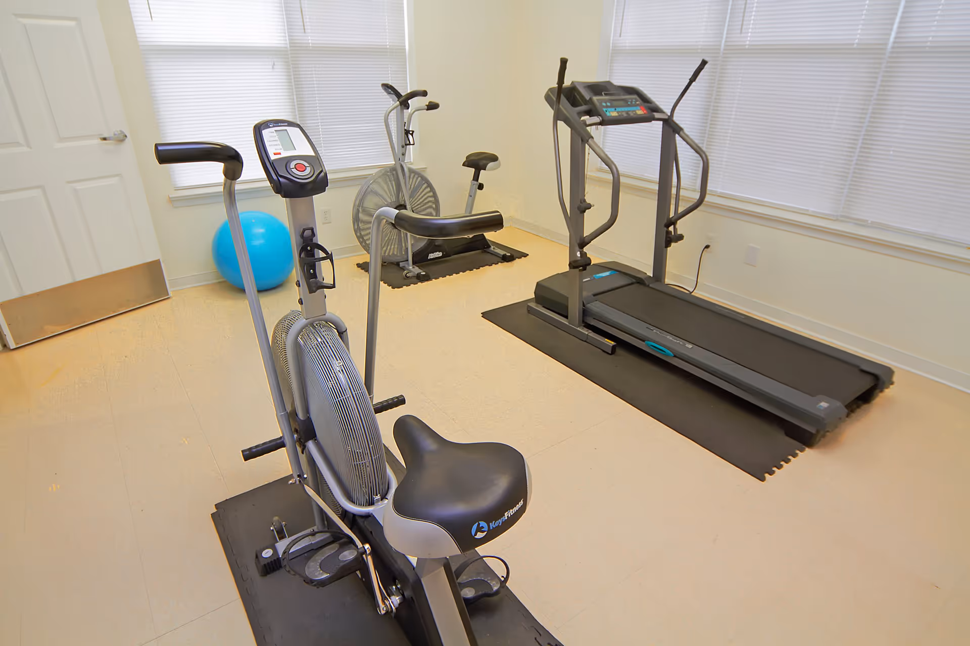 Small exercise room with stationary bikes, a treadmill, and a blue exercise ball by bright windows.