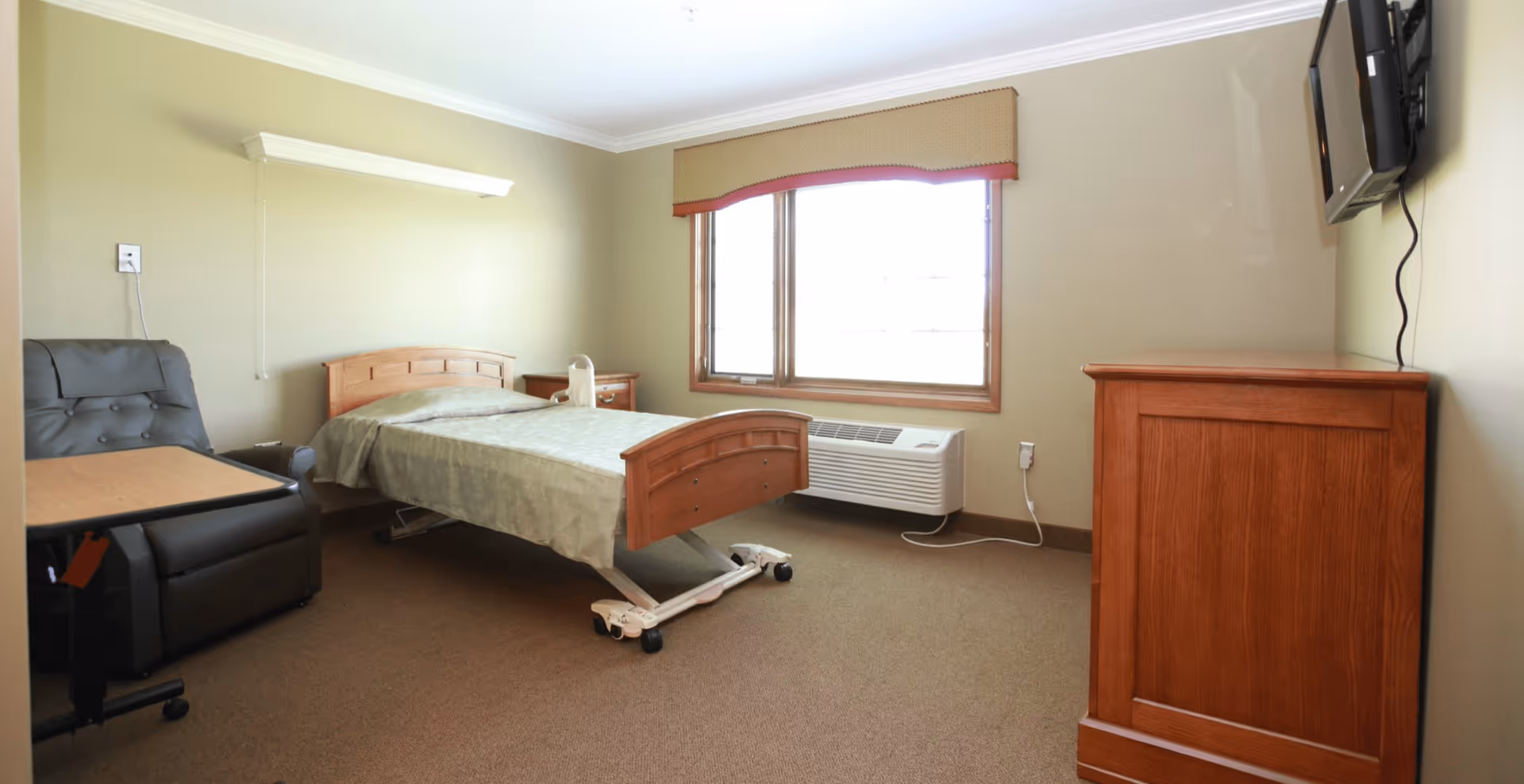 A simple, clean bedroom in a senior living facility with a single adjustable bed, a padded recliner chair with a small table, a wooden dresser, a wall-mounted TV, and a large window with a beige valance allowing natural light to fill the room.