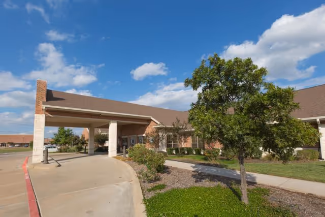 Front entrance of a brick healthcare building with a covered drive-up canopy, sidewalk, and landscaped trees under a blue sky.