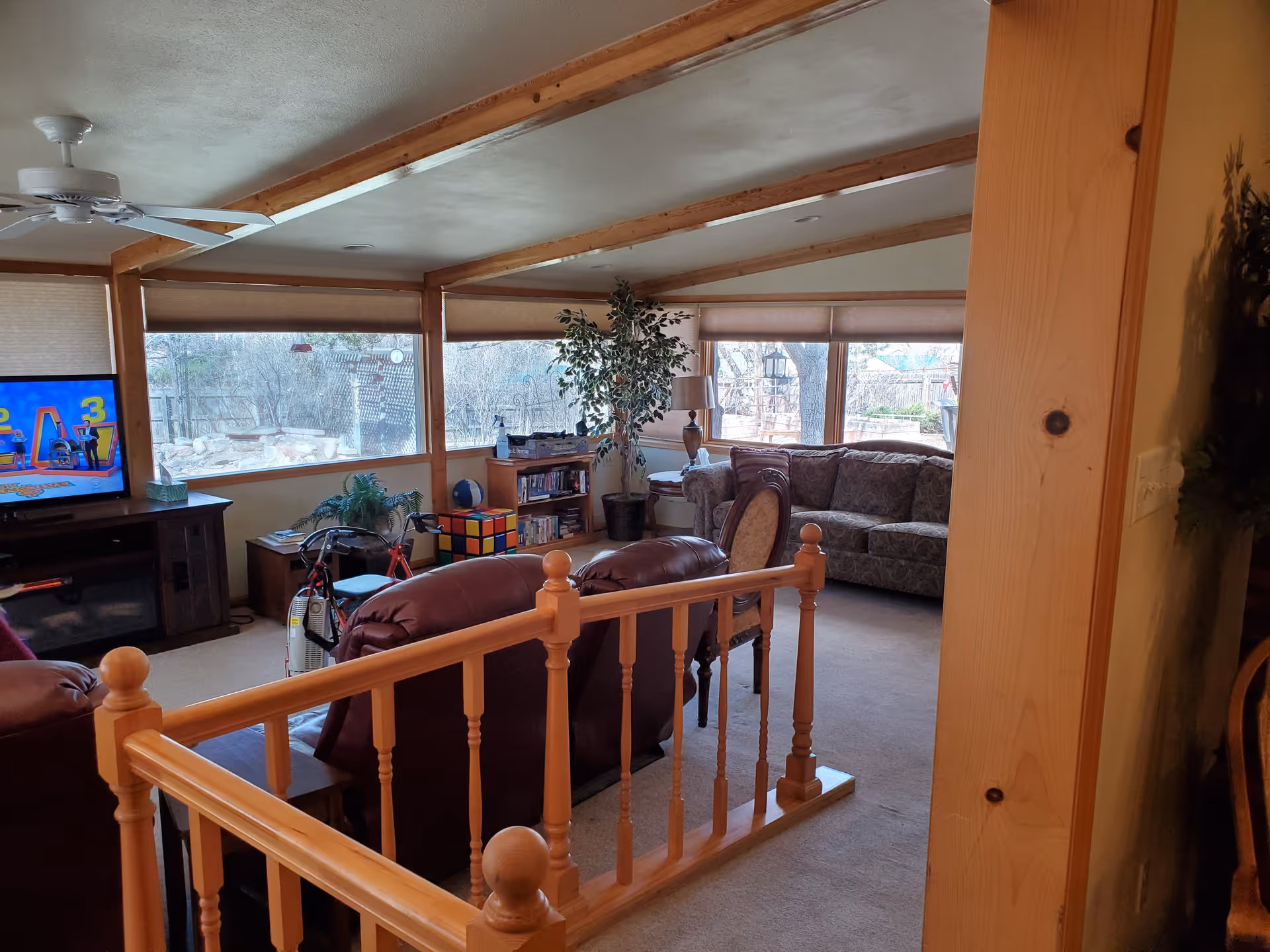 A cozy living room area with large windows showing an outdoor view. The room features a ceiling fan, wooden beams, a TV on a stand, a large Rubik's cube, a walker, a bookshelf, a potted plant, a lamp, a patterned sofa, and two leather recliners separated by a wooden railing.