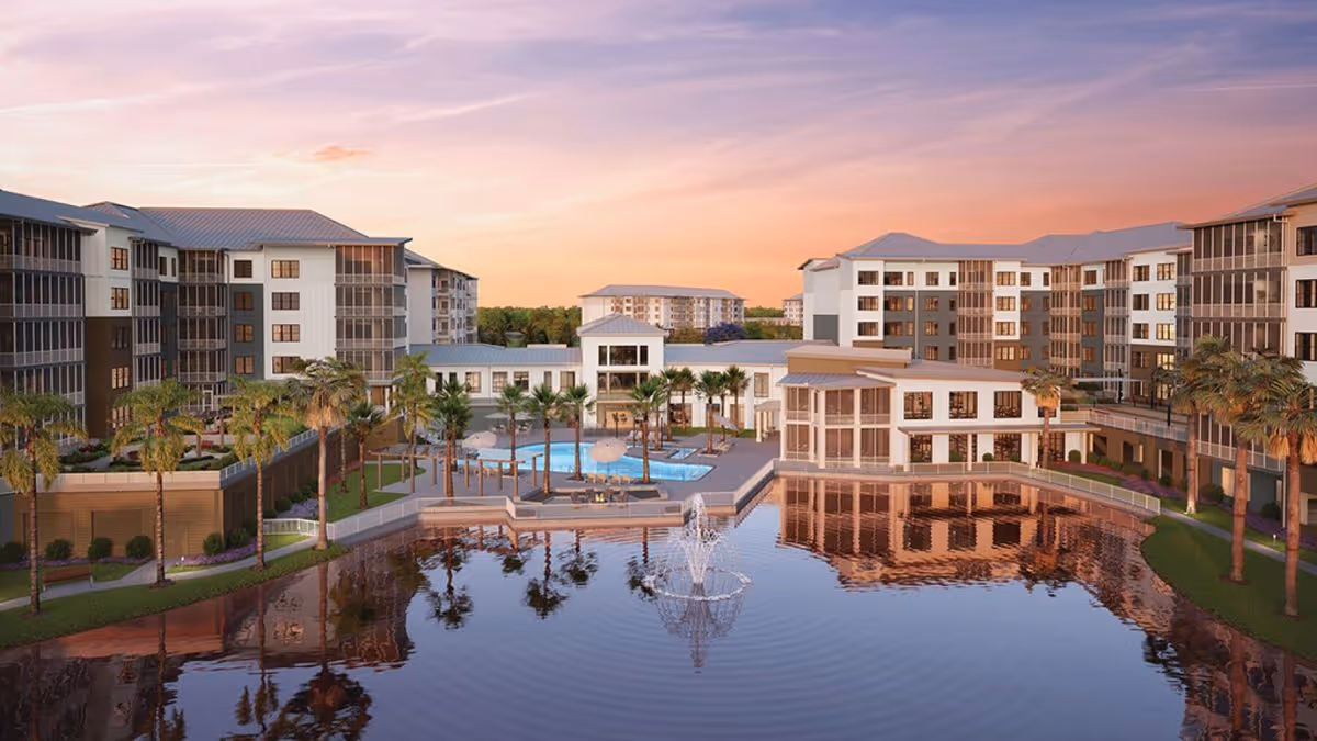 A senior living complex surrounding a central lake with a fountain, pool, palm trees, and multi-story buildings at sunset.