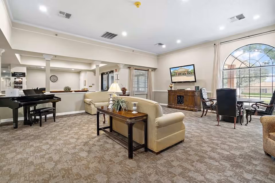 A spacious senior living facility common area with beige sofas, a wooden coffee table with plants and candles, a black grand piano with a matching bench, a large arched window with curtains, and a flat-screen TV mounted on the wall above a wooden cabinet. The room has carpeted flooring and neutral-colored walls with ceiling lights.