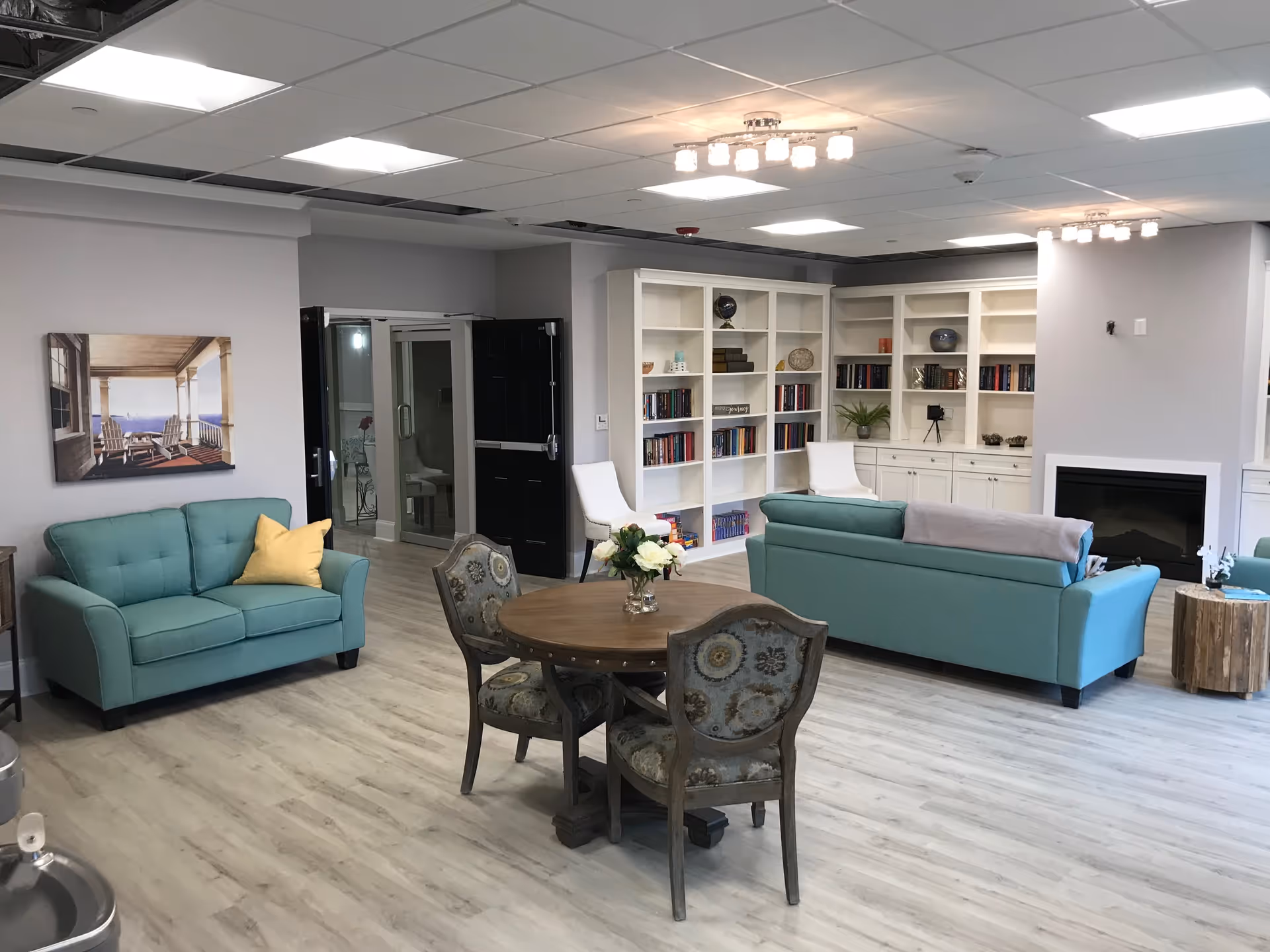 Bright communal living room with teal sofas, a round wooden table and chairs, built-in bookshelves and a fireplace.