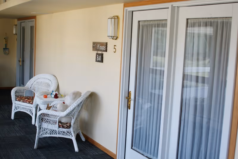 A hallway in a senior living facility with two white wicker chairs and a small table between them. The chairs have patterned cushions and a stuffed animal is placed on one chair. There is a door with white framed glass panels and curtains on the right side. A wall-mounted light fixture and a wooden sign with the names 'Koops' and 'Bill' and the number 5 are visible on the wall.