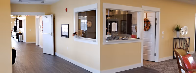 Interior view of a hallway in an assisted living community with beige walls and wood flooring. There is a reception window with a 'welcome' sign, a door decorated with a fall wreath, and a seating area with chairs and a small table with a plant and decorative item.