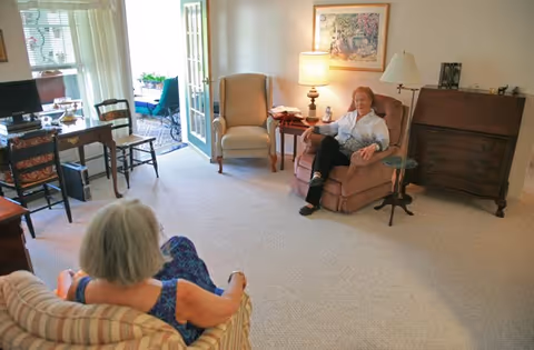 Two elderly women sitting and conversing in a well-lit living room with beige carpet, armchairs, a wooden desk with a computer, a side table with a lamp, and a painting on the wall. A door opens to an outdoor patio area with chairs.