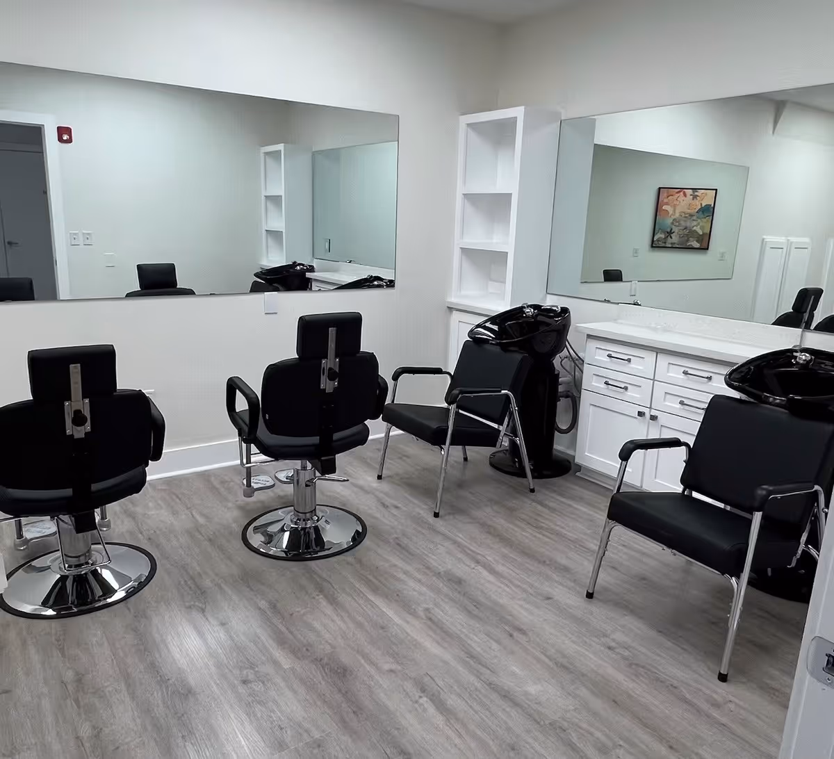 Interior of a salon room with three black salon chairs, two with attached hair washing sinks, large mirrors on the walls, white cabinetry with drawers, and light wood flooring.