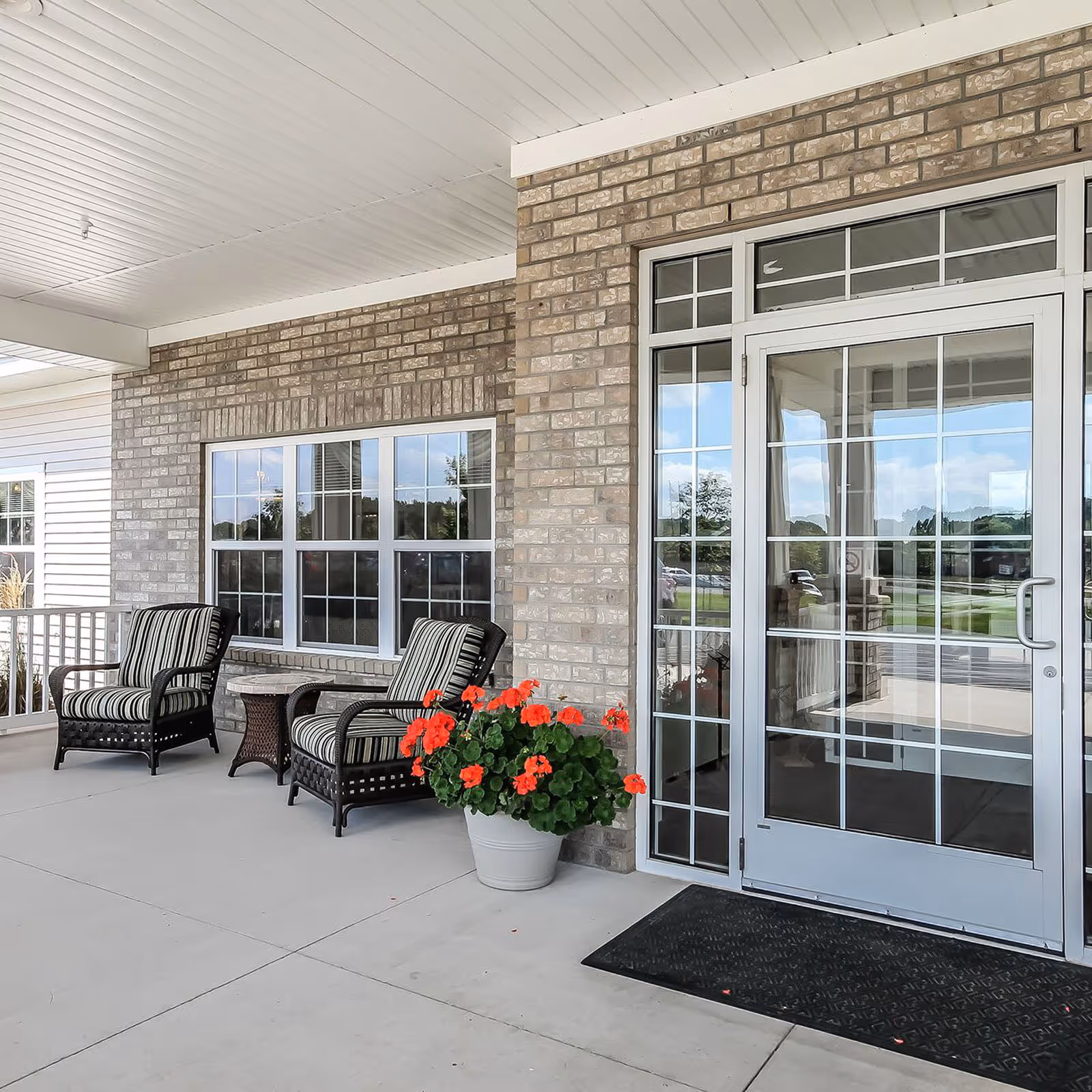 Covered outdoor patio area with two cushioned wicker chairs, a small wicker table, a potted plant with red flowers, and a glass door entrance with window panels on a brick wall.