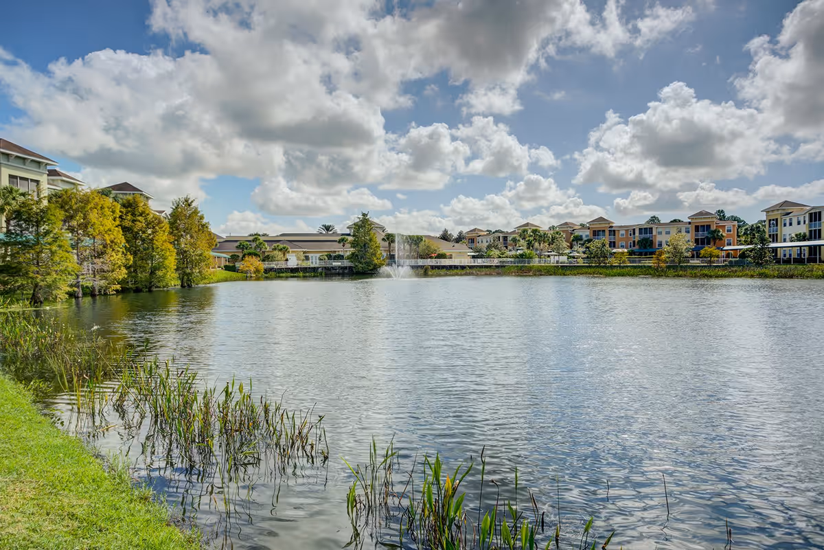 A scenic view of a large pond with a fountain in the center, surrounded by green grass and trees. Residential buildings are visible in the background under a partly cloudy sky.