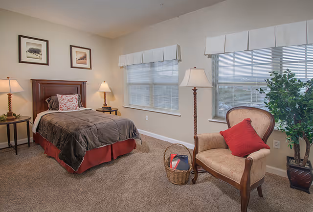 Furnished bedroom with a twin bed, two bedside tables and lamps, an upholstered armchair with a red pillow, a wicker basket, and two windows.