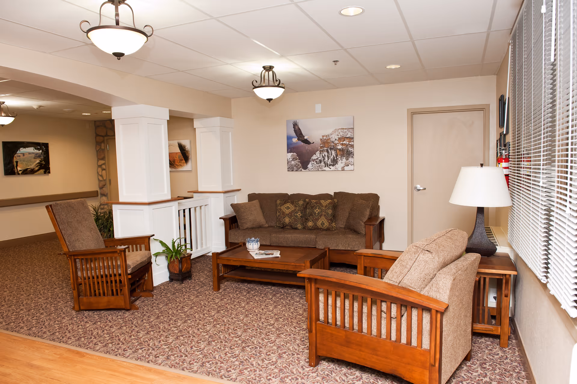 A cozy living room area in a senior care facility featuring a brown sofa with patterned cushions, two wooden armchairs with beige cushions, a wooden coffee table with a small decorative item and a magazine, a side table with a lamp, and wall art depicting an eagle flying over a canyon. The room has beige walls, carpeted flooring, ceiling lights, and a window with blinds.