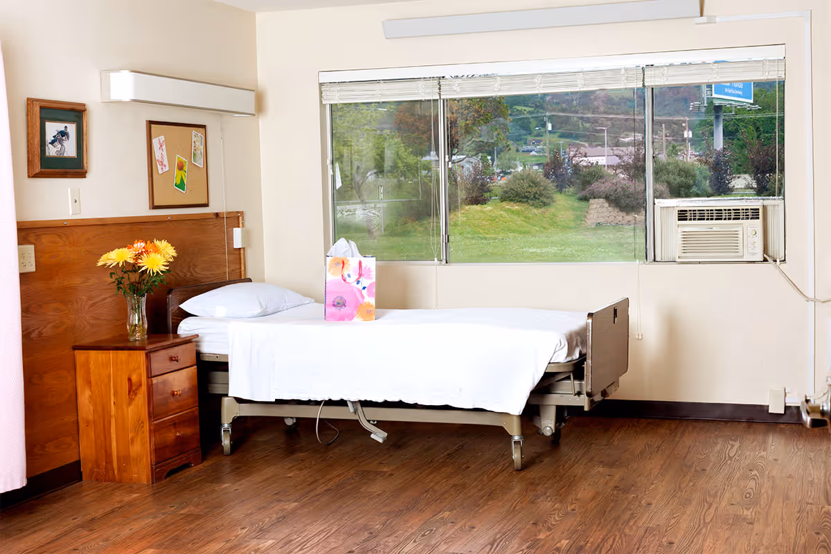 A nursing center bedroom with a single hospital-style bed covered with white linens, a wooden nightstand with a vase of yellow flowers, a bulletin board with pinned cards, a framed picture on the wall, large windows showing greenery outside, and a window air conditioning unit.