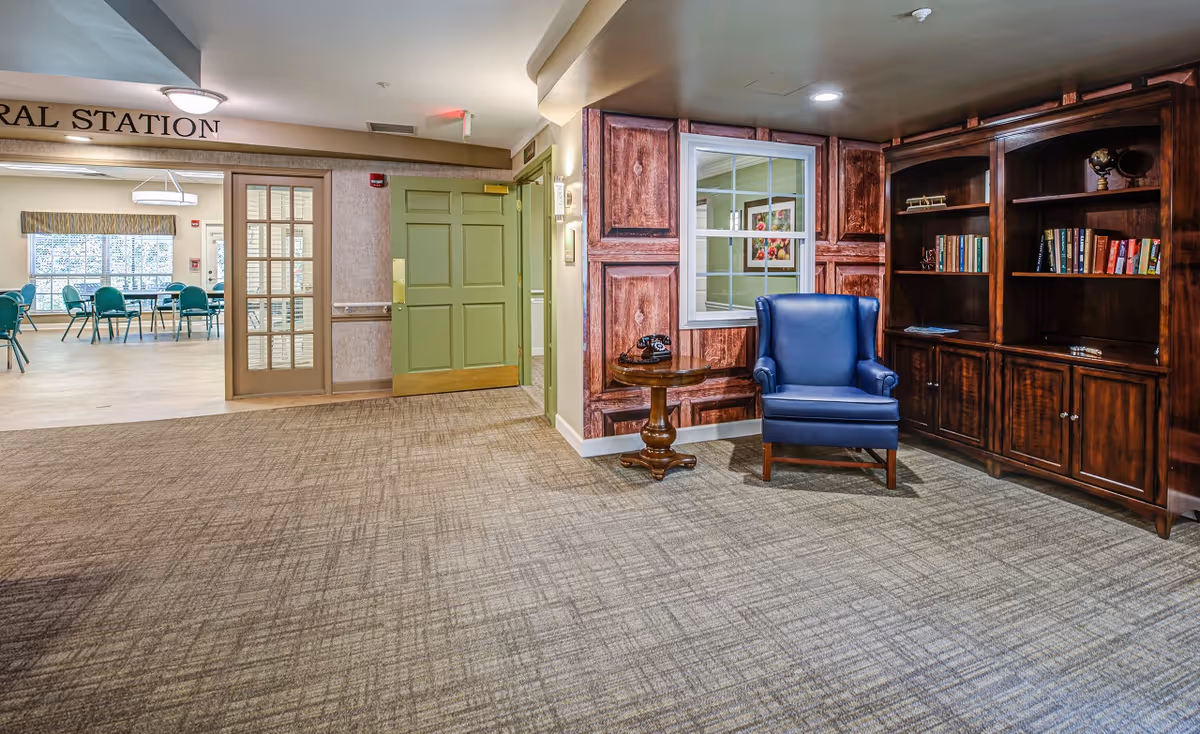 Interior view of a senior living facility showing a carpeted hallway with a blue armchair next to a wooden side table holding a vintage telephone. There is a large wooden bookshelf filled with books on the right. A green door is open leading to a dining area with tables and green chairs. The wall has a window and decorative wood paneling.