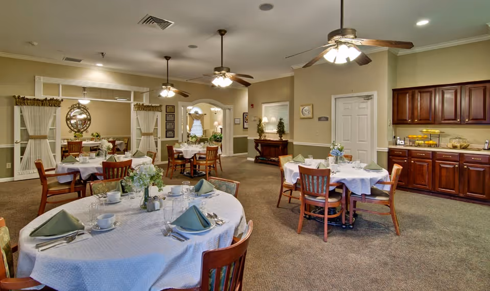 A dining room in a senior living facility with round tables covered in white tablecloths, each set with plates, cups, silverware, and green folded napkins. The room has wooden chairs, ceiling fans with lights, a sideboard with cabinets and food items, and decorative elements including flowers and framed pictures on the walls.
