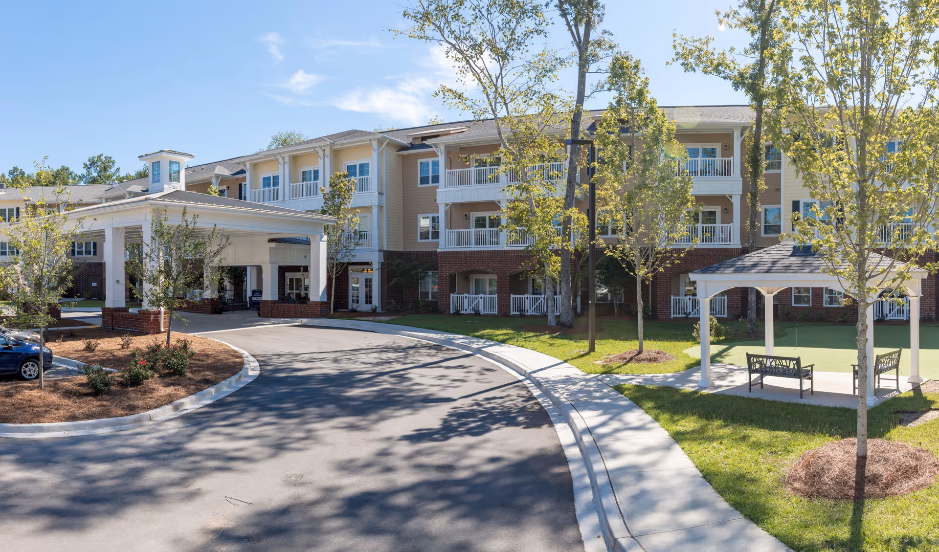 Exterior view of a senior living facility named Harmony at Wescott, showing a three-story building with balconies, a covered entrance driveway, landscaped trees, and a small gazebo with benches on a sunny day.