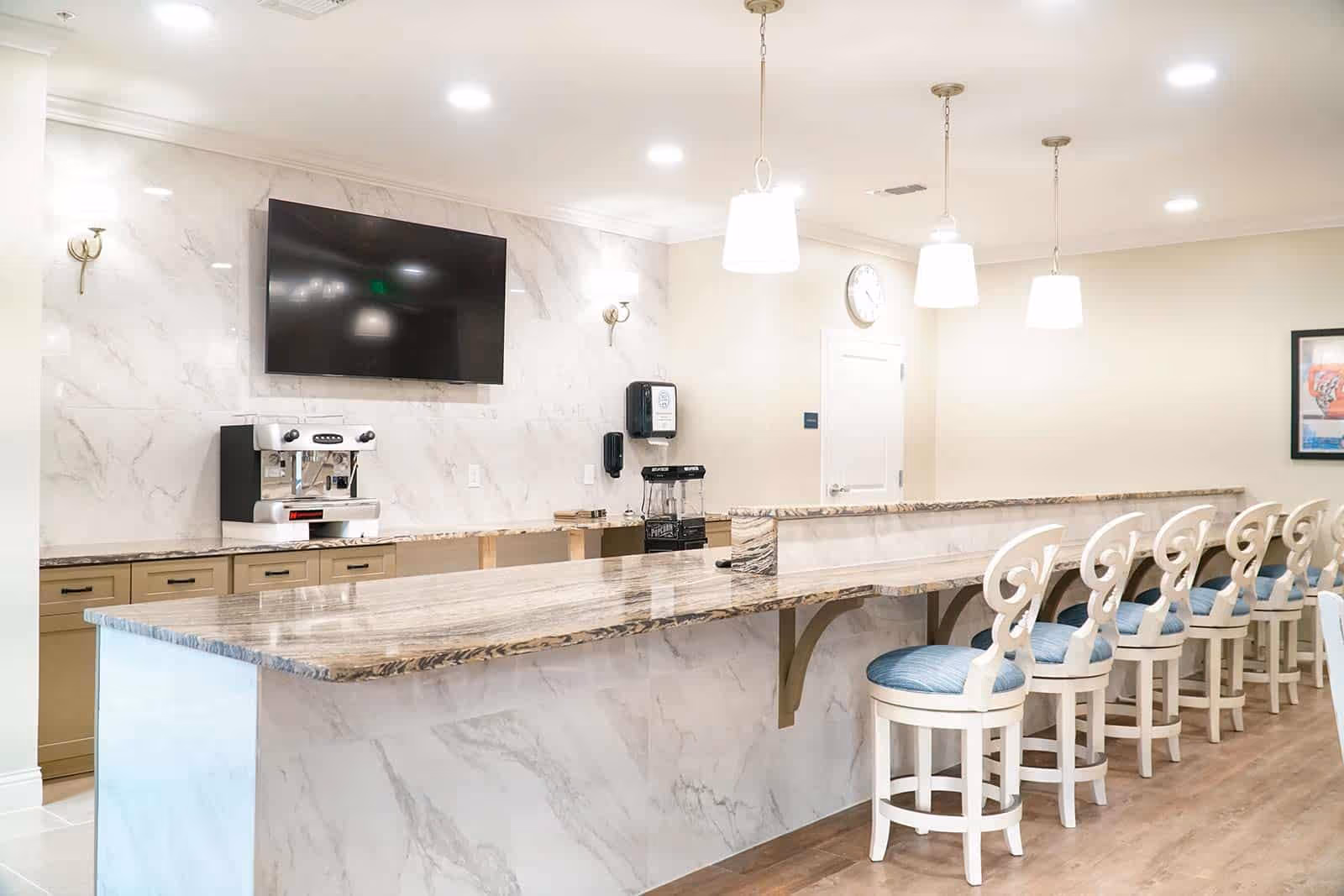 Bright communal bar area with a long marble countertop, a row of white bar stools with blue cushions, a coffee machine, and a wall-mounted TV.