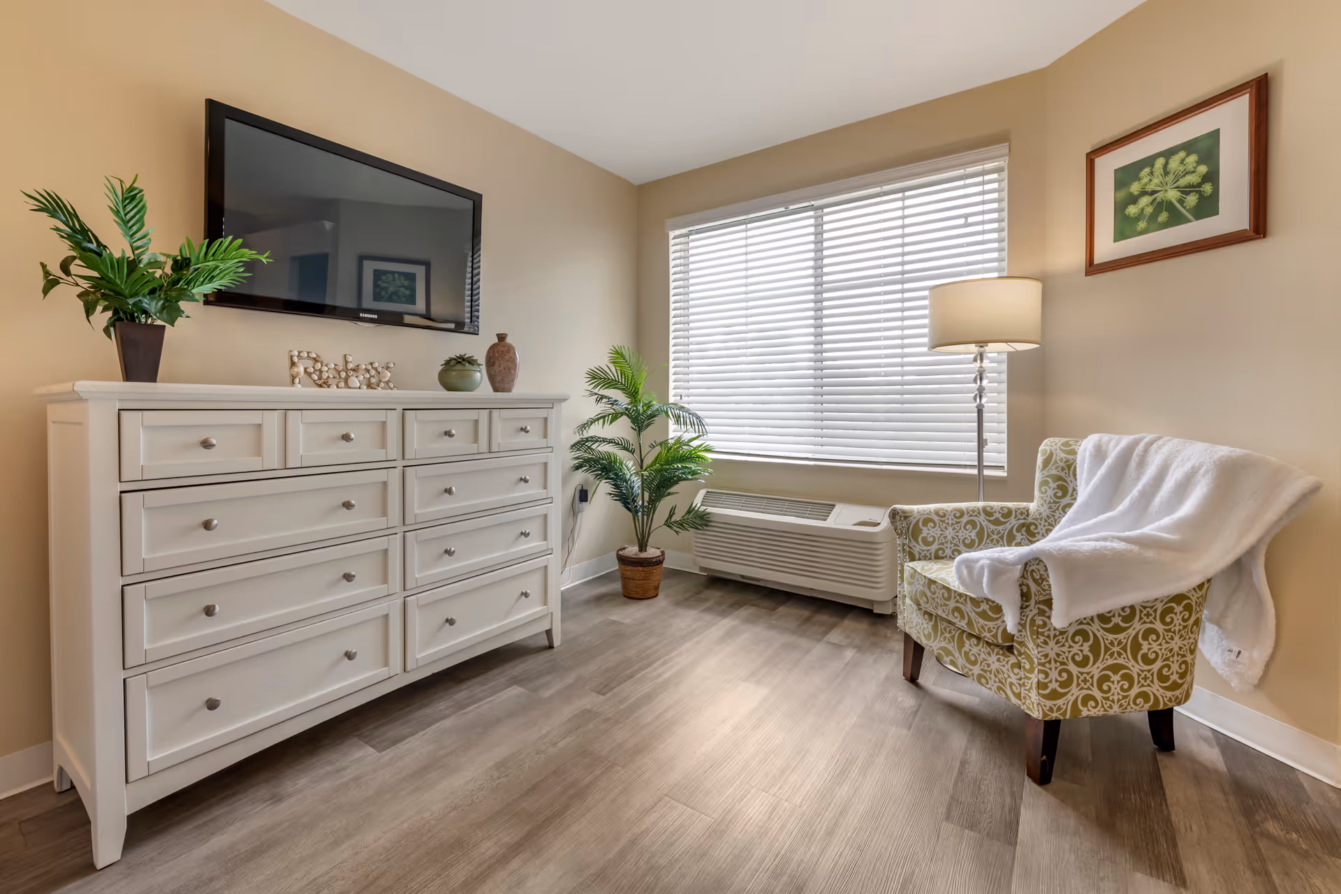 A cozy living room area with a white dresser topped with decorative items including a plant and a 'Relax' sign. A flat-screen TV is mounted on the beige wall above the dresser. Next to a large window with white blinds, there is a green patterned armchair draped with a white blanket and a tall floor lamp. A potted plant is placed near the window, and the floor is covered with light wood flooring.