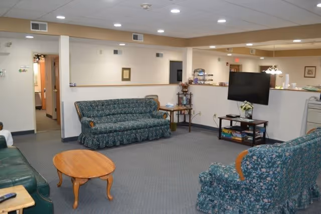 A common area in an assisted living facility featuring multiple upholstered sofas with floral patterns, a wooden coffee table, a flat-screen TV on a stand with shelves holding books and magazines, and a small side table with decorative items. The room has carpeted flooring, white walls, and ceiling lights. There is an open doorway leading to another room.