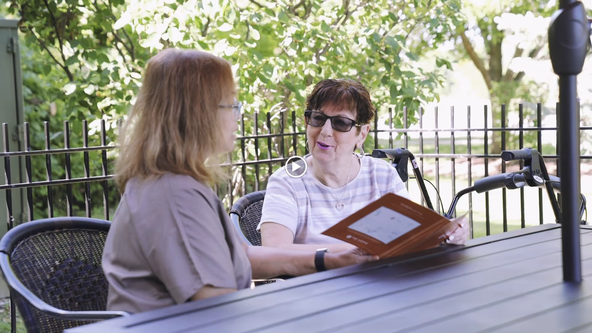 Two women sitting at a black outdoor table under an umbrella, engaged in conversation. One woman is holding a booklet or menu. They are surrounded by greenery and a black metal fence is visible in the background.