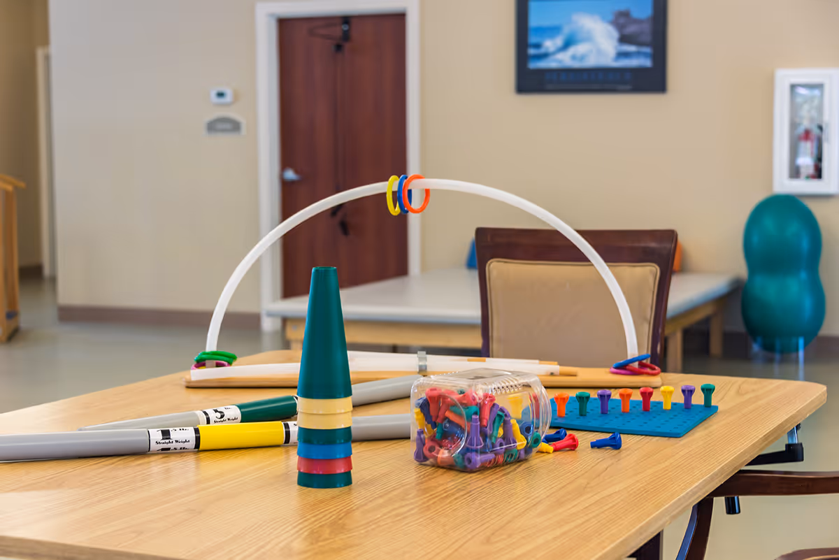 A table in a senior activity room holding colorful therapy toys—stacking cones, pegboard pegs, rings, and plastic tubes—with chairs and an exercise ball in the background.