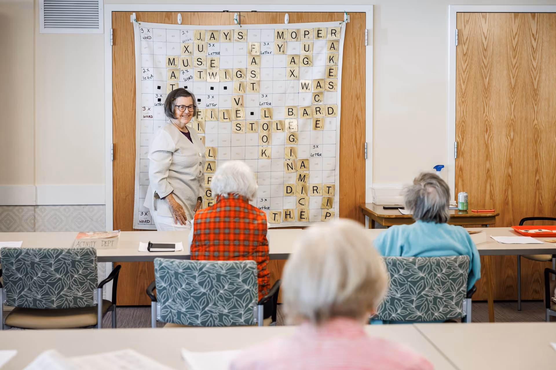 An elderly woman standing and smiling in front of a large wall-mounted Scrabble board, with three elderly people seated at tables facing her in a communal activity room.