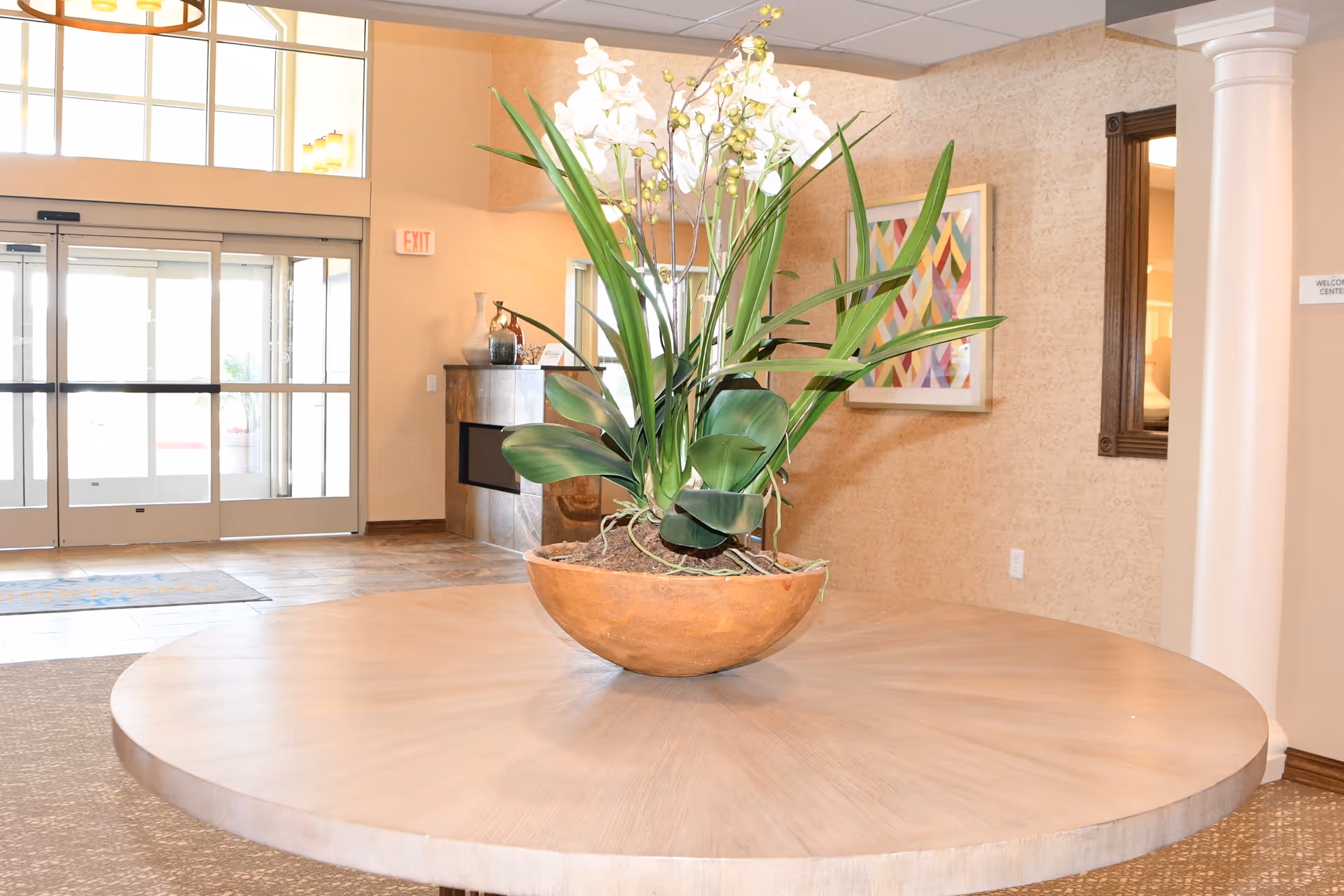 A large round wooden table with a decorative plant in a brown pot placed in the center, situated in a bright lobby area with glass entrance doors, beige walls, a framed colorful geometric artwork, and a white column.
