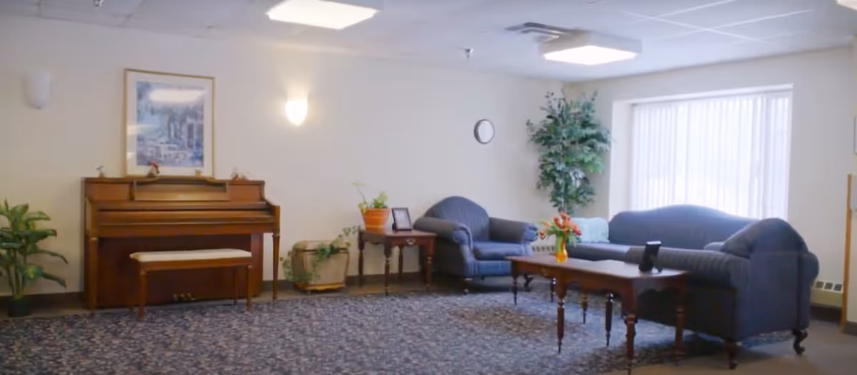 A cozy living room area in Nature's Point Assisted Living featuring a wooden piano with a bench, a framed picture above it, several potted plants, a blue upholstered armchair and sofa, a wooden coffee table with a vase of flowers, and a large window with vertical blinds letting in natural light.