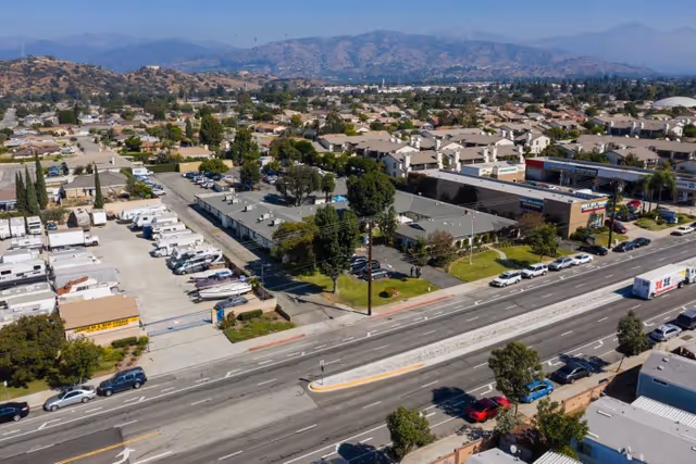 Aerial view of a suburban area showing a single-story building with a parking lot, surrounded by trees and residential houses. There are several cars parked along the street and in the parking lot. Mountains are visible in the background under a clear sky.