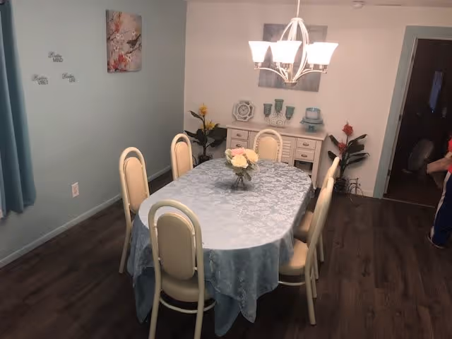 A dining room with a rectangular table covered by a light blue tablecloth and surrounded by six cream-colored chairs. A vase with flowers is placed in the center of the table. The room has wooden flooring, light blue walls, and a chandelier with five lights hanging above the table. There is a white sideboard against the wall with decorative items and two plants on either side. A painting hangs above the sideboard, and a small wall decoration with words is visible on the left wall.