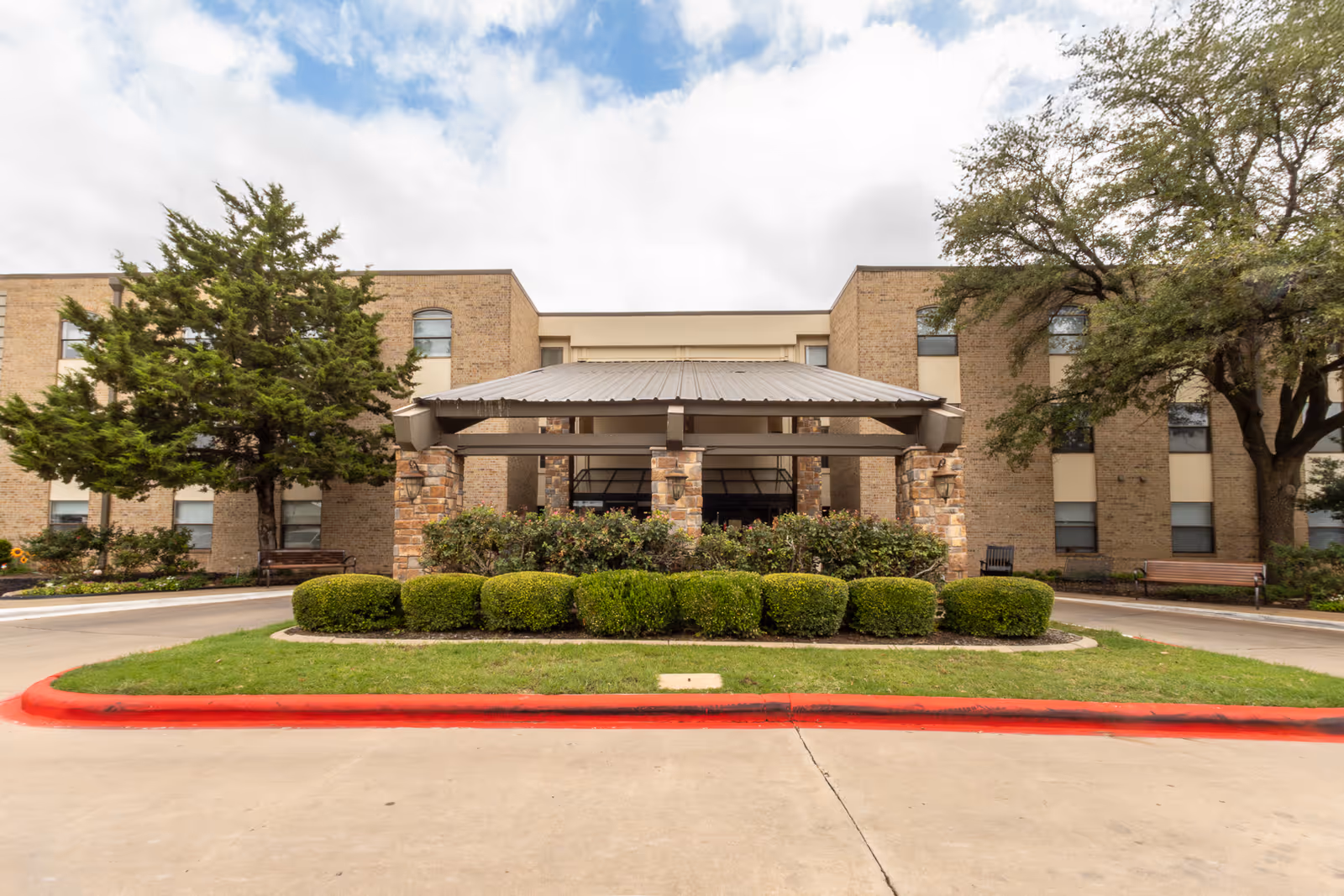 Front entrance of a three-story brick senior living building with a covered portico, trimmed shrubs and trees.