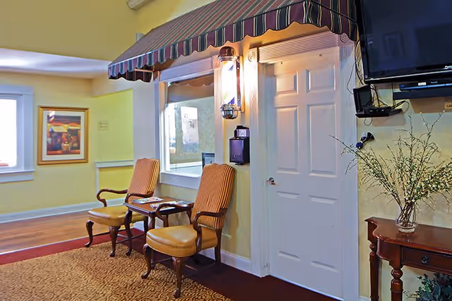 A cozy interior corner of a senior living facility with two cushioned wooden chairs and a small table between them under a striped awning. There is a white door next to the chairs, a wall-mounted lantern light, a mounted television, and a small wooden table with a vase of flowers. The walls are painted yellow and there is a framed picture on the far wall.
