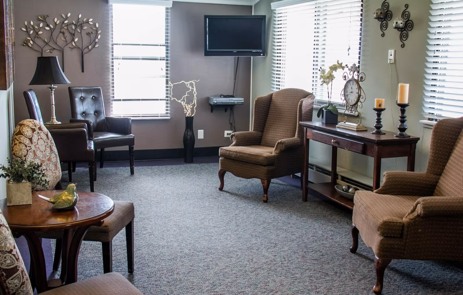 A cozy living room area with several upholstered chairs, a small wooden table with a decorative bird figurine, a wooden console table with candles, a clock, and a potted plant. The room has carpeted flooring, two windows with blinds, a wall-mounted TV, and decorative wall art.