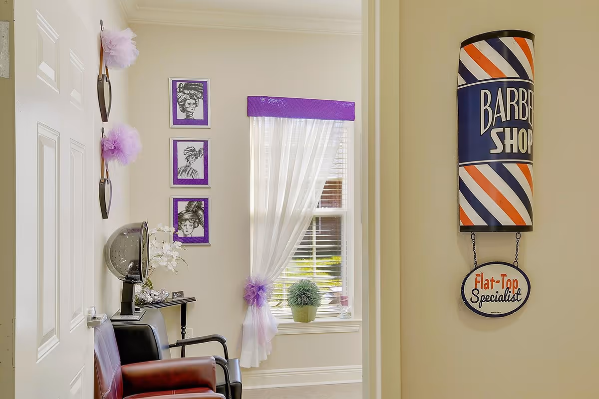 Interior view of a senior living facility's barber shop area with a vintage barber pole sign on the wall, a window with sheer white curtains tied with purple ribbons, three framed black and white portraits with purple borders, a black and red salon chair, and a hair dryer hood.