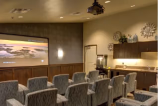 A small theater room with rows of upholstered chairs facing a large screen on the wall. The room has warm lighting, wood paneling on the lower half of the walls, and cabinets with decorative items on the right side.