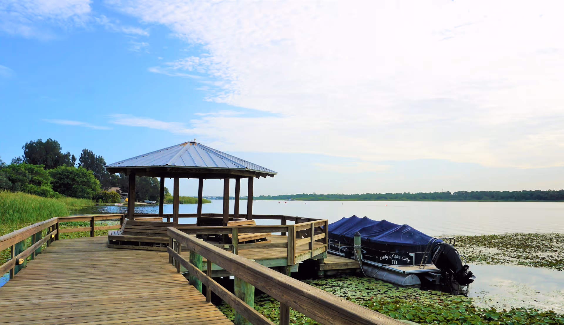 A wooden dock extending over a lake with a covered gazebo seating area. A pontoon boat named 'Lady of the Lake III' is moored alongside the dock. The lake is surrounded by greenery and trees under a partly cloudy sky.
