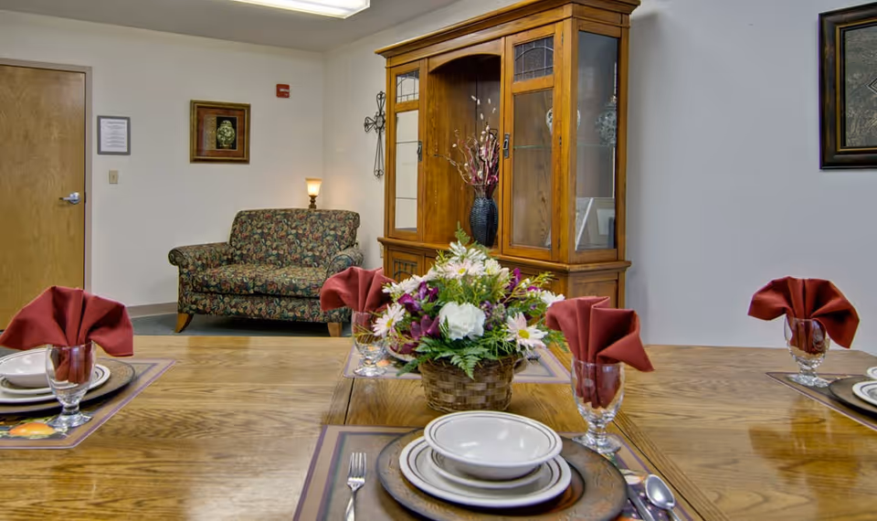 A dining area with a wooden table set with plates, bowls, glasses, and folded red napkins. In the center of the table is a floral arrangement in a basket. In the background, there is a floral-patterned loveseat, a wooden cabinet with glass doors displaying decorative items, a wall lamp, and framed artwork on the walls.